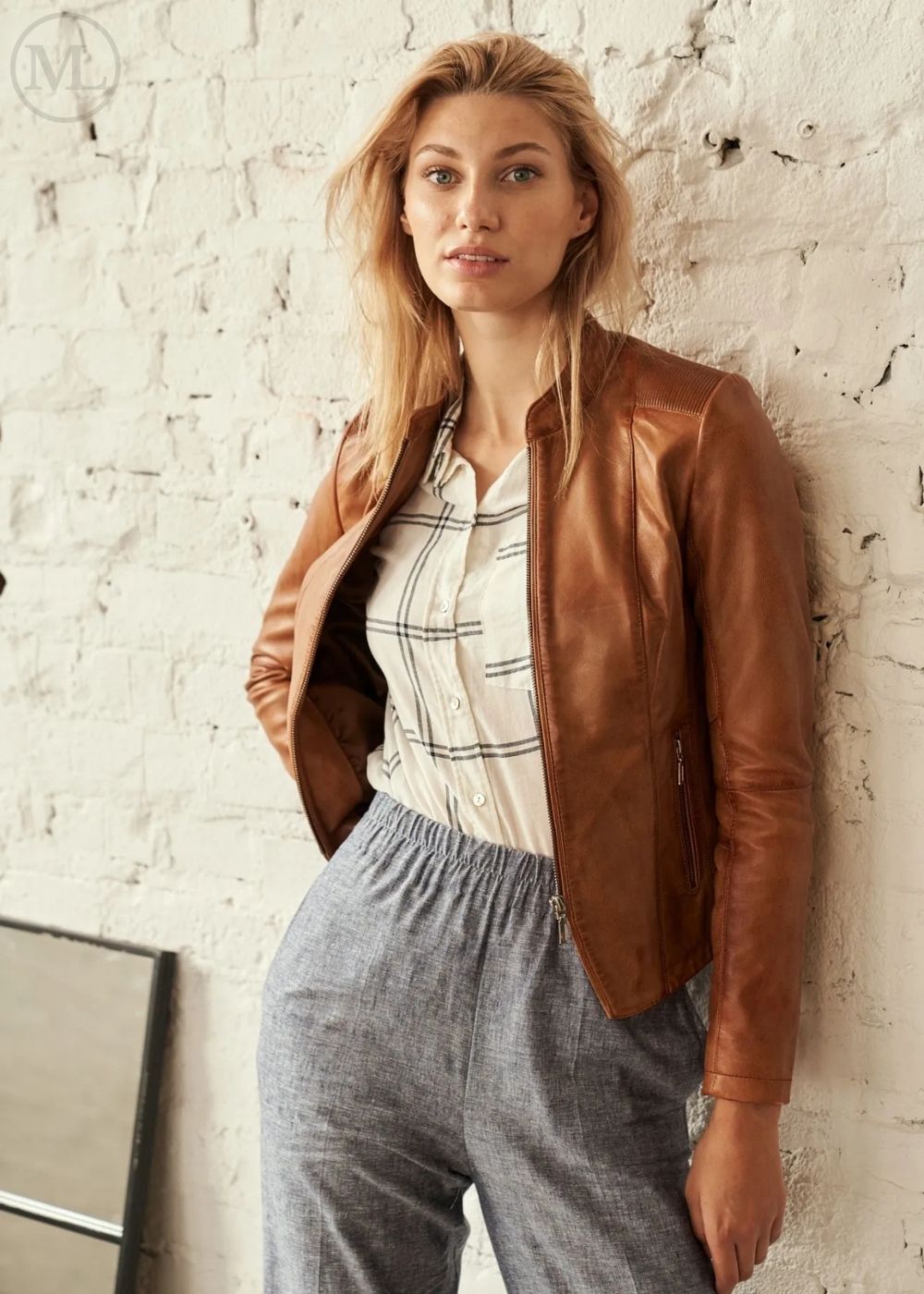 Woman wearing a cognac brown leather jacket over a plaid shirt and gray pants against a textured white wall.