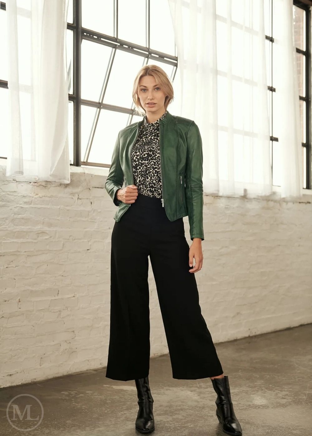 Woman wearing a green jacket, black patterned top, and black wide-leg pants in a bright room with large windows.