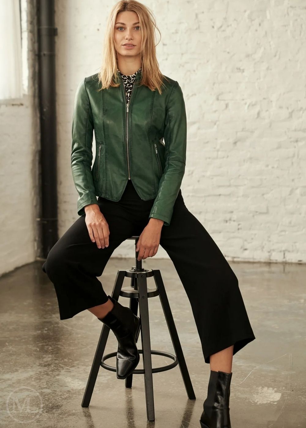 Woman wearing a green leather jacket sitting on a stool against a white brick wall.