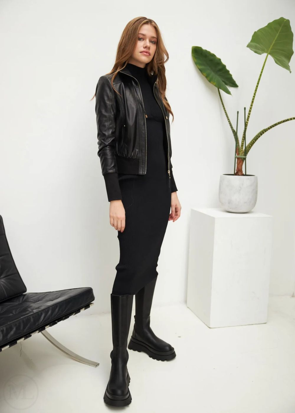 Woman in a black outfit with a leather bomber jacket standing in a minimalistic room.