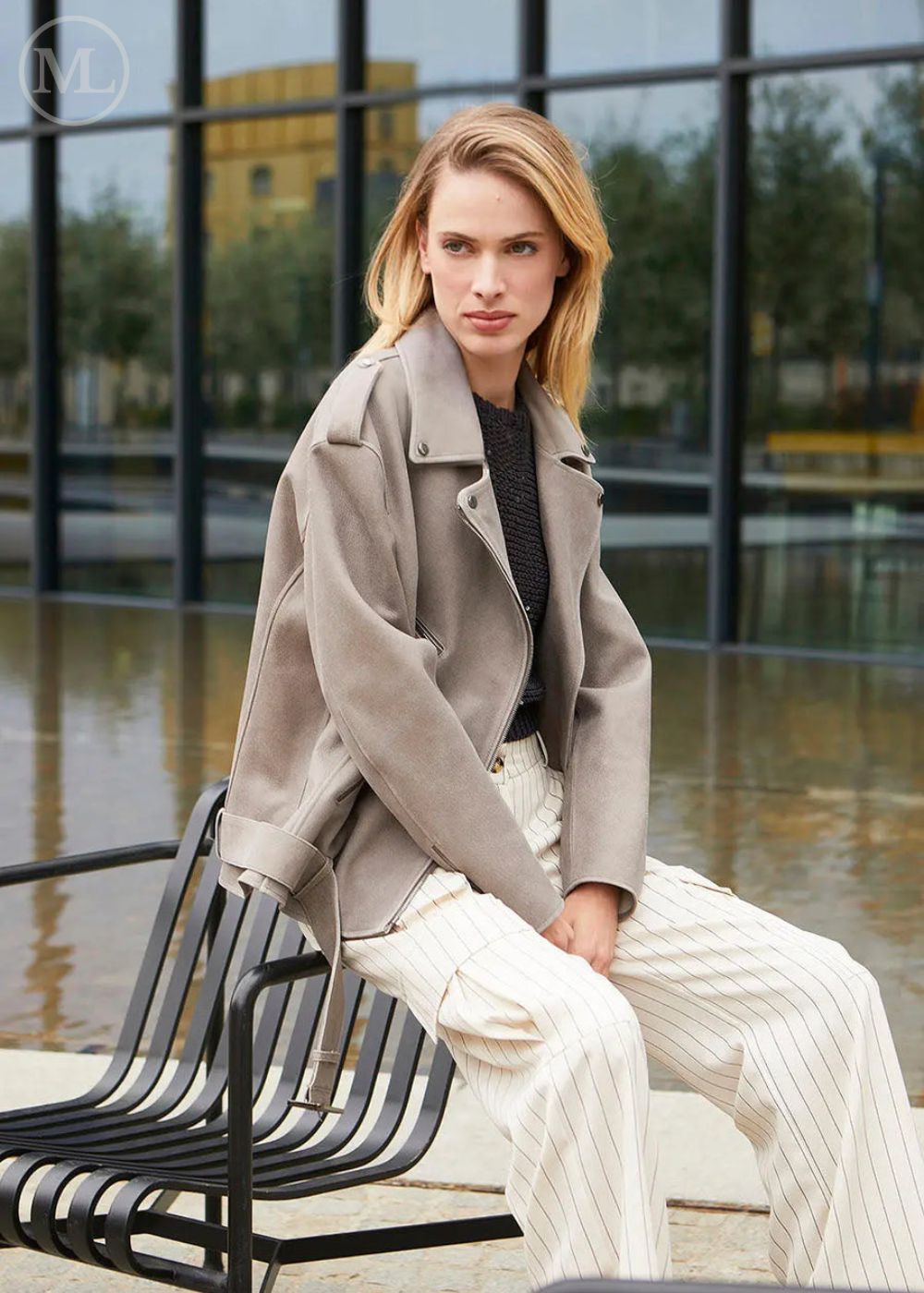 Femme assise sur un banc portant une veste de motard surdimensionnée en cuir nubuck gris avec ceinture et un pantalon blanc dans un cadre urbain.