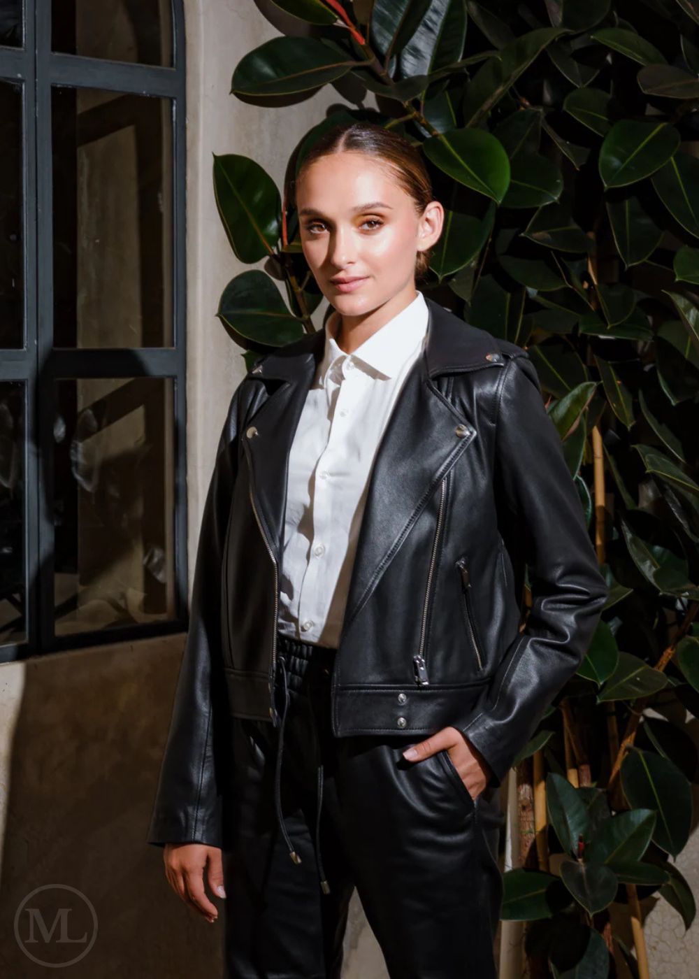 Women wearing a black leather biker jacket and white shirt standing against a plant backdrop