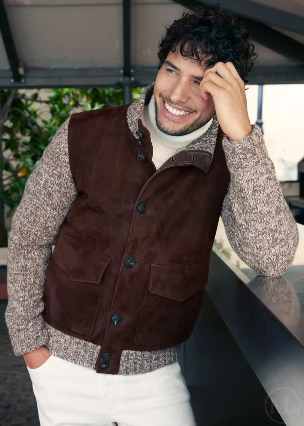 Man wearing a dark brown jacket over a patterned sweater with a blurred indoor background