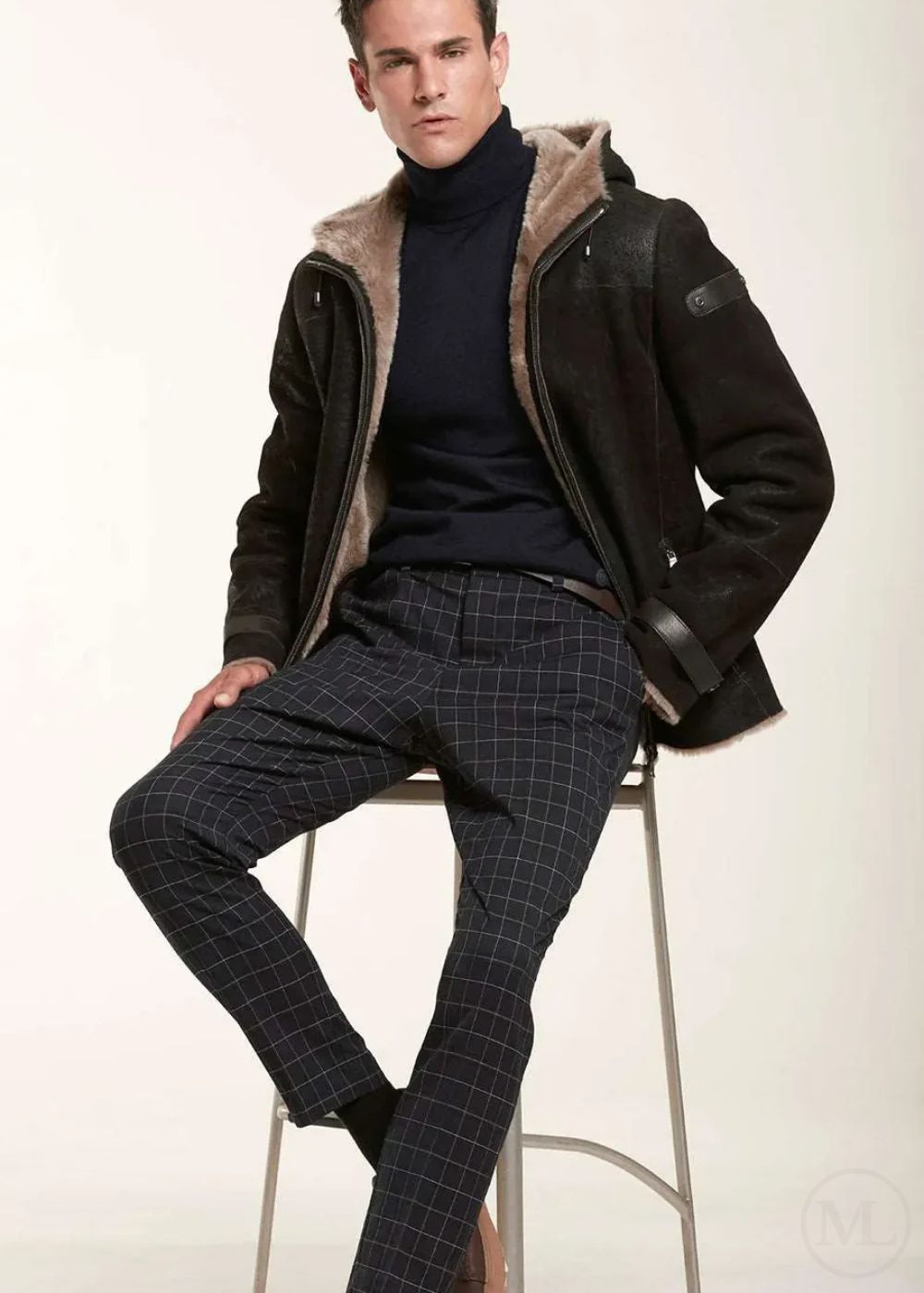 Man wearing a dark brown jacket with a hood, navy turtleneck, and checkered pants on a white background
