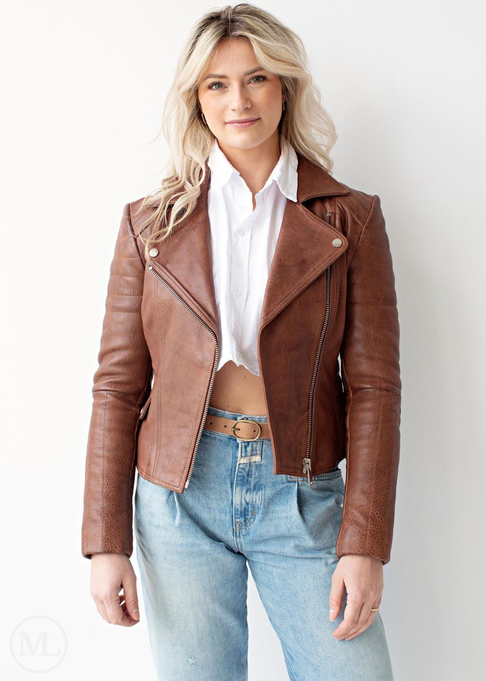 Woman wearing a cognac brown leather jacket, white shirt, and blue jeans against a plain background