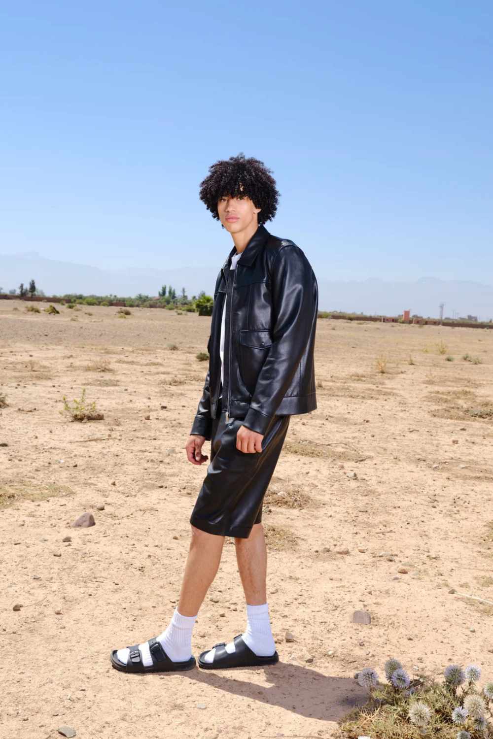 a young boy standing in desert wearing a Designer men’s khaki leather trucker jacket with flap chest pockets, and welt side pockets and leather shorts
