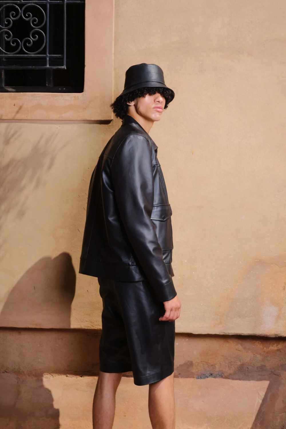 a young boy standing in side post wearing a Designer men’s khaki leather trucker jacket with flap chest pockets, and welt side pockets pairing it with leather shorts and leather bucket hat
