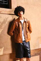 a young boy with curly hair standing again a wall,wearing a Designer men’s camel color leather trucker jacket with flap chest pockets, and welt side pockets, pairing it wih leather shorts