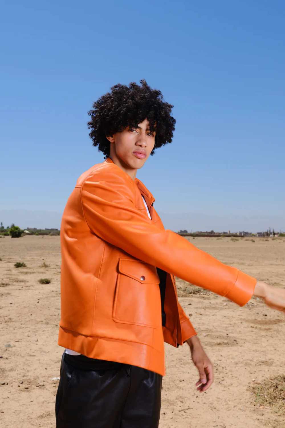 a young boy with curly hair standing in a side pose wearing a Designer men’s orange leather trucker jacket with flap chest pockets, and welt side pockets in a desert and clear blue sky
