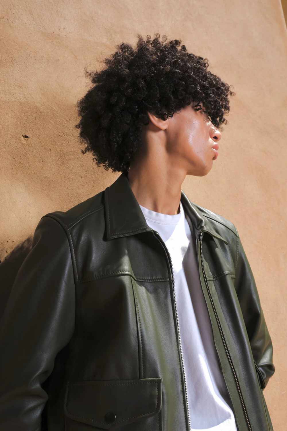a young boy resting on beige wall wearing a Designer men’s khaki leather trucker jacket with flap chest pockets, and welt side pockets, close shot
