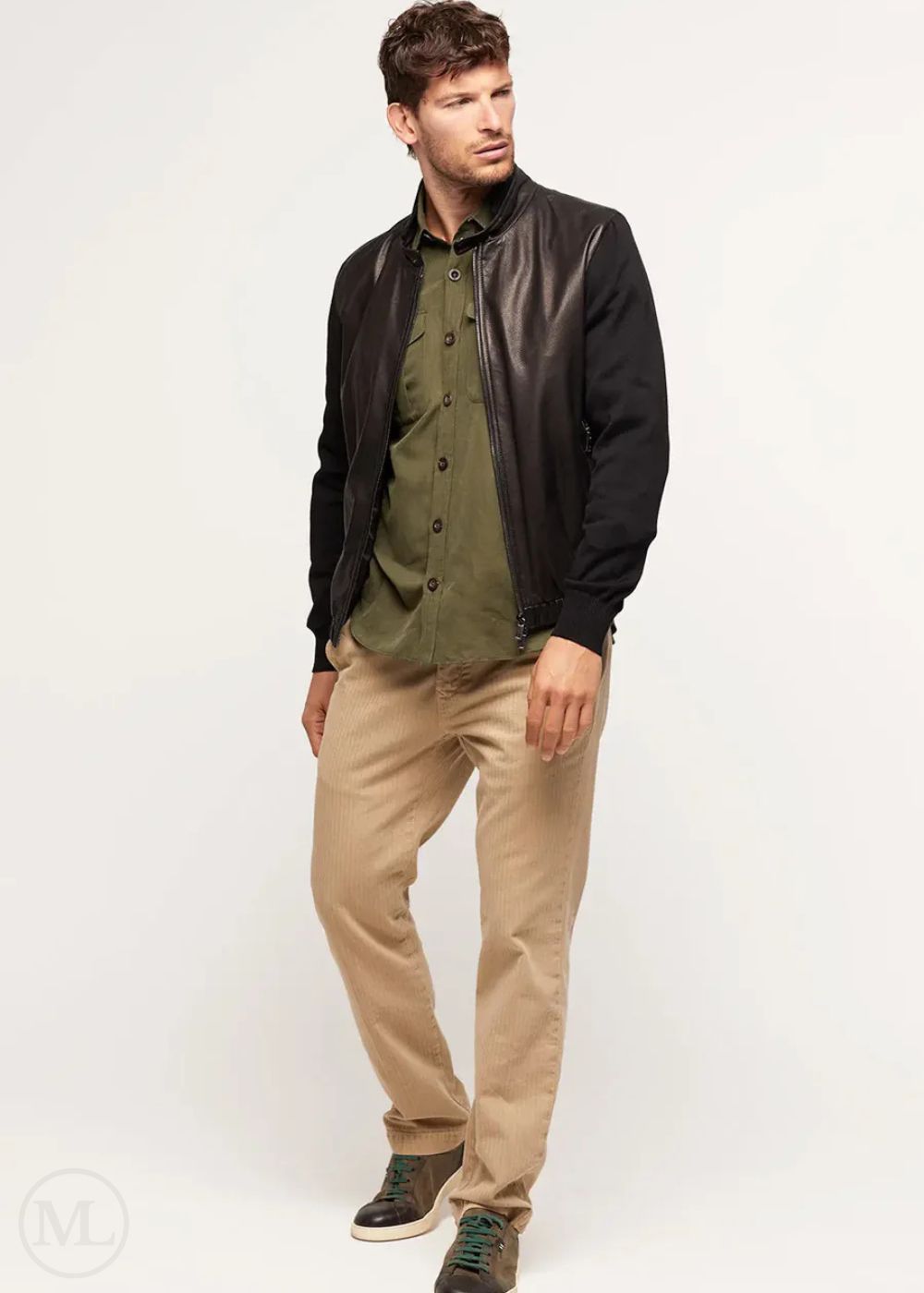 Man wearing a black leather bomber jacket, green shirt, and beige pants on a white background