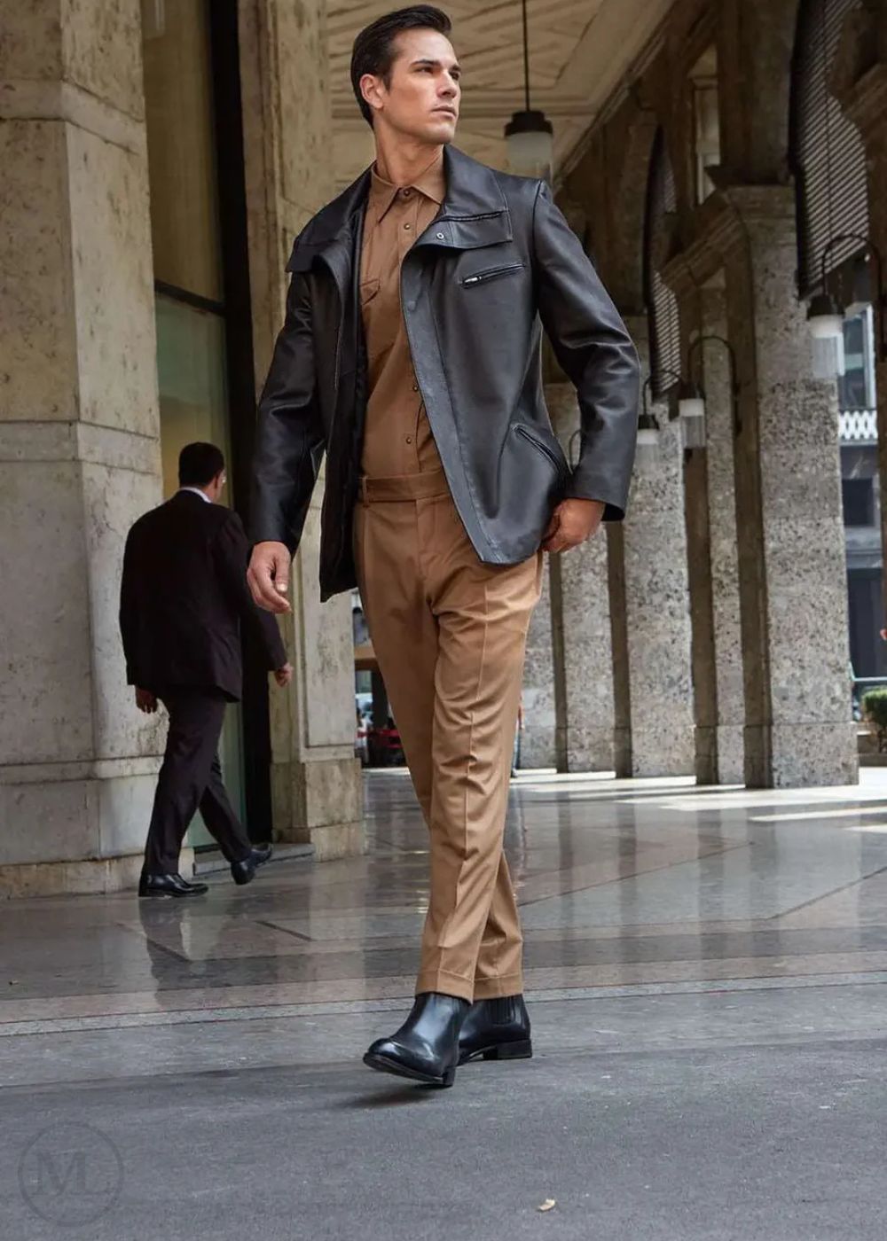Man in a stylish outfit with a black jacket and brown pants walking in an urban setting.