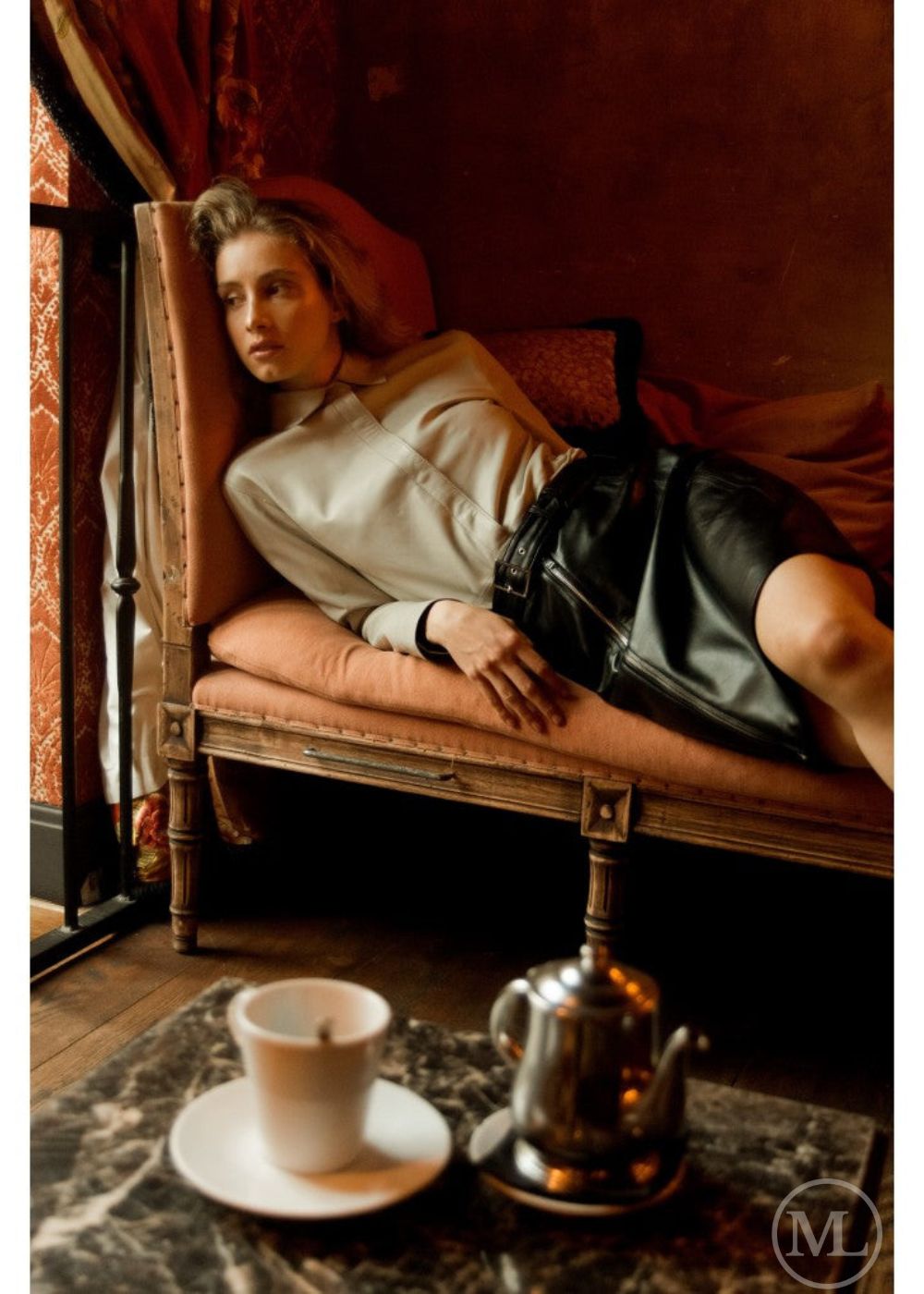 Woman wearing ivory leather shirt lying on a bed with a cup and teapot on a table in front of her.