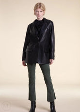 Woman wearing a black leather blazer jacket, black top, green pants, and black boots on a beige background