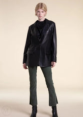 Woman wearing a black leather blazer jacket, black top, green pants, and black boots on a beige background