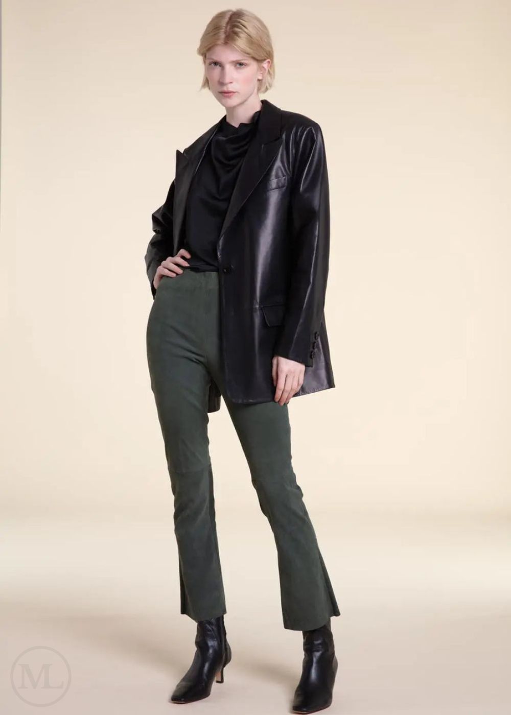 Woman wearing a black leather blazer jacket, black top, and green pants on a beige background