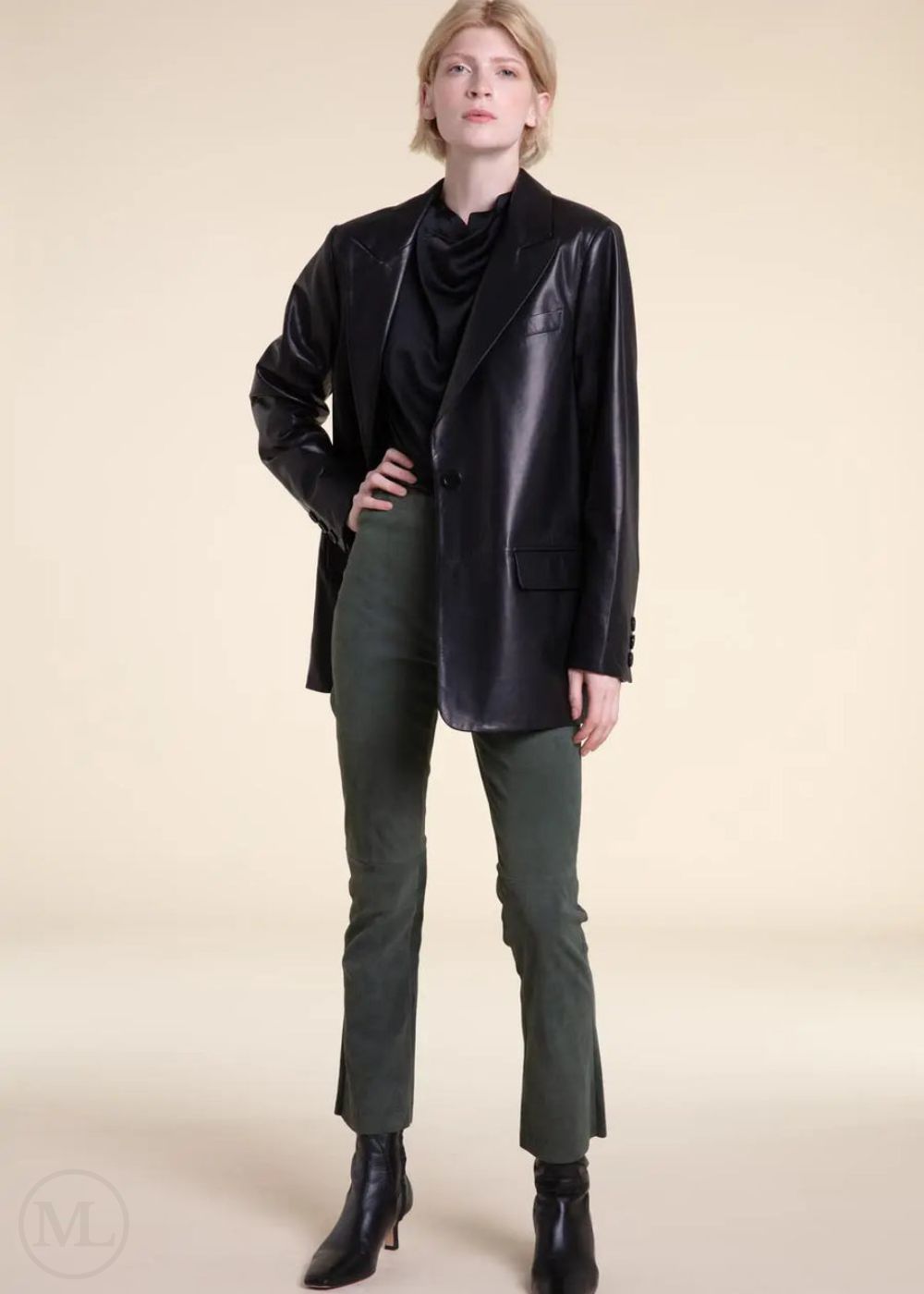 Woman wearing a black leather blazer jacket, black top, and green pants on a beige background