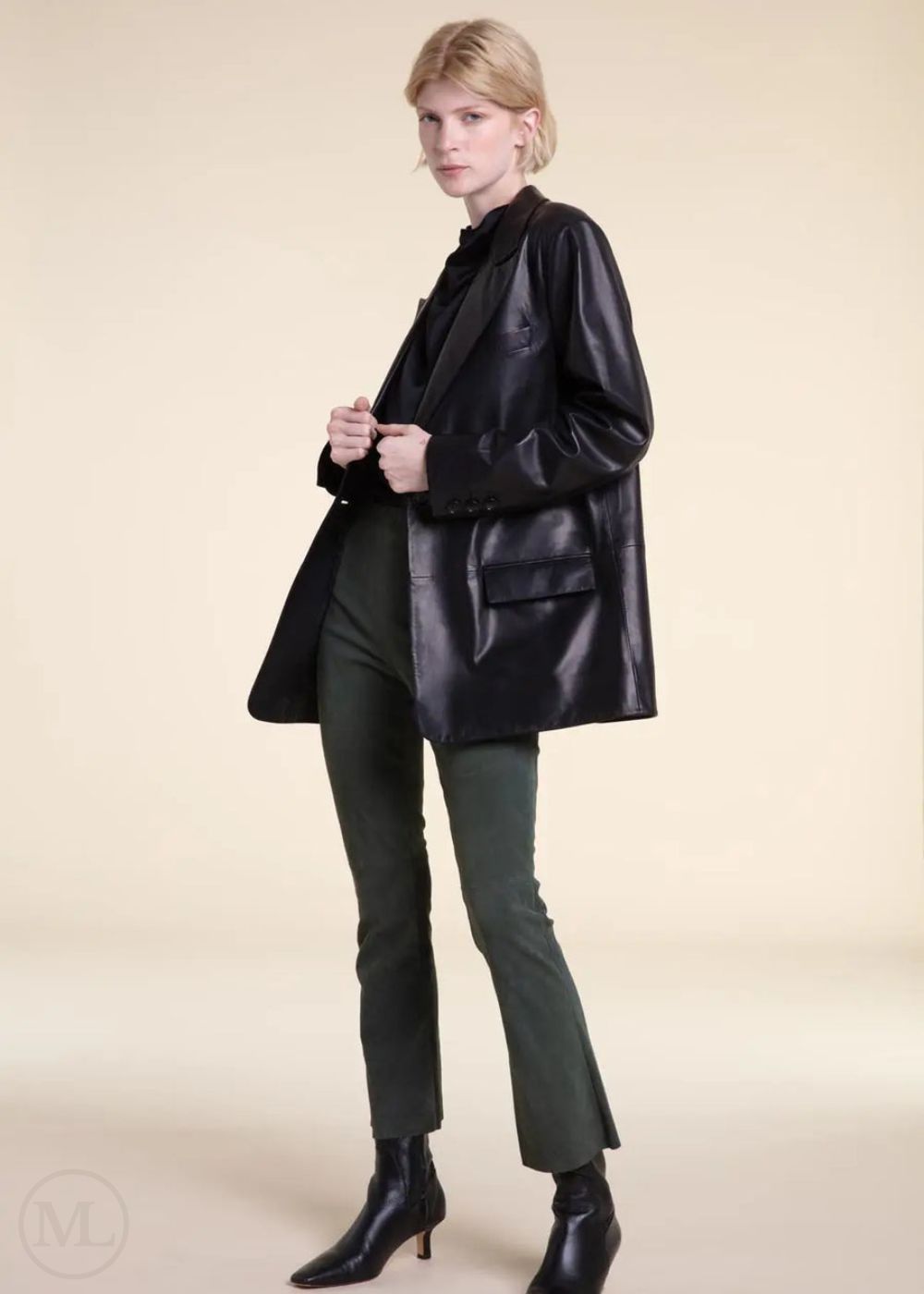 Woman wearing a black leather blazer jacket, green pants, and black boots on a beige background