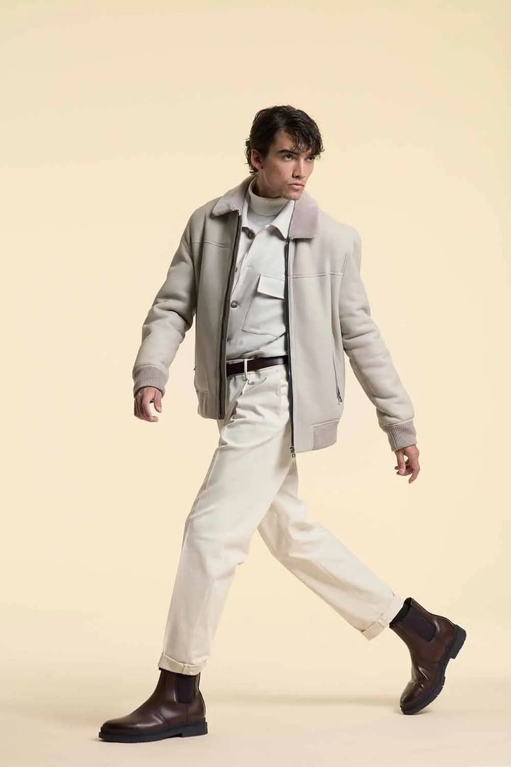 Man wearing a white shearling leather jacket, white shirt, beige pants, and brown boots on a beige background