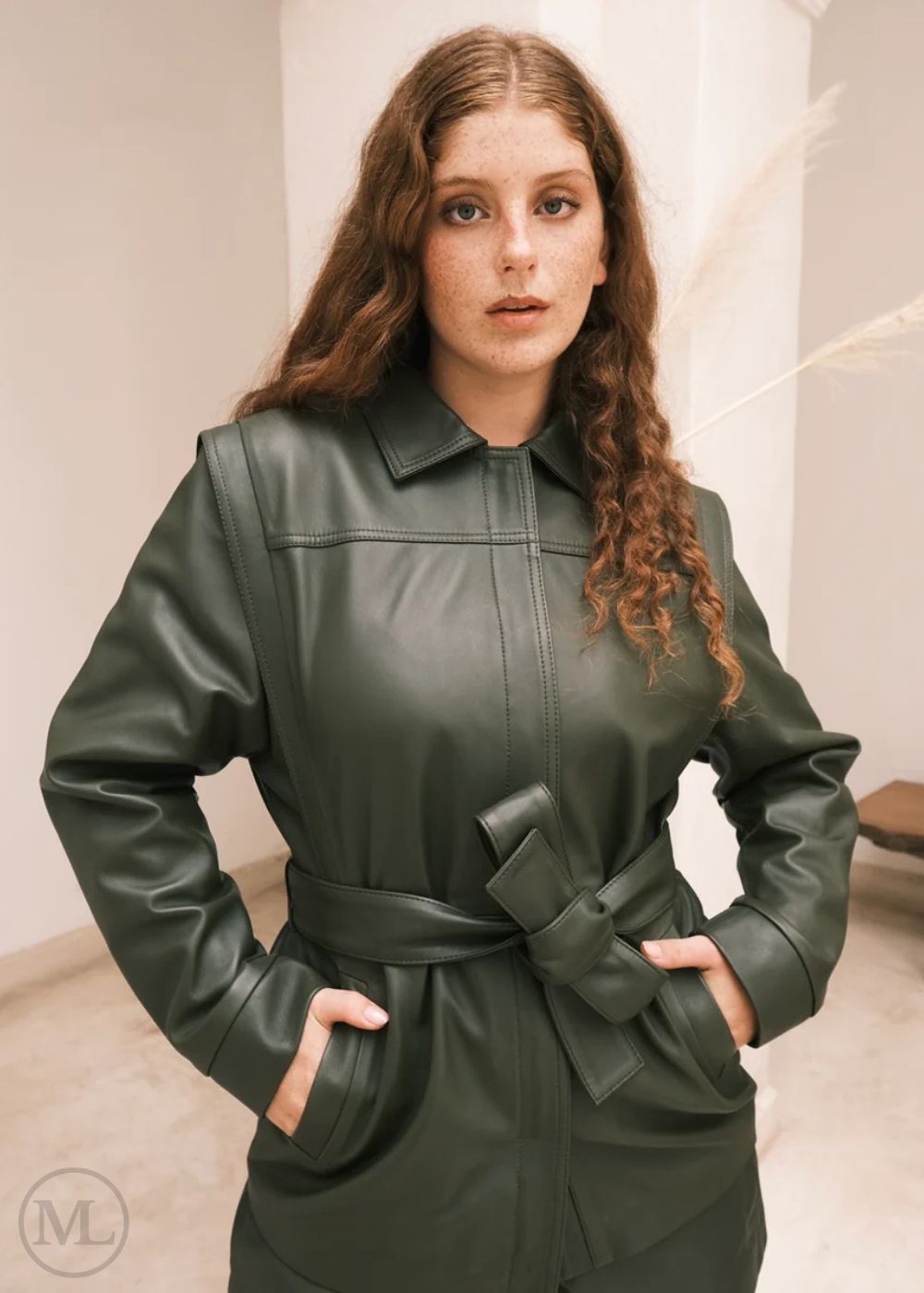 Femme portant une veste en cuir vert avec une ceinture, debout sur un fond neutre.