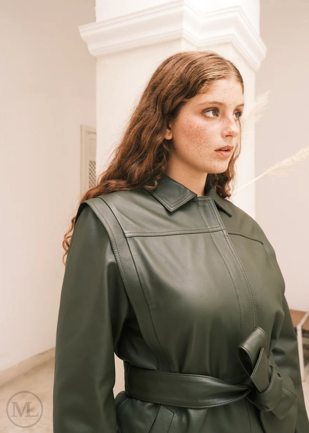 Femme portant une veste en cuir vert avec une ceinture, debout sur un fond neutre.
