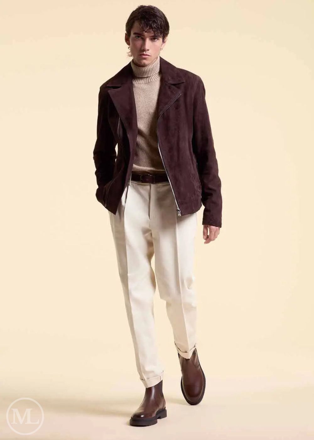 Man wearing a dark brown suede jacket, beige sweater, white pants, and brown shoes on a beige background