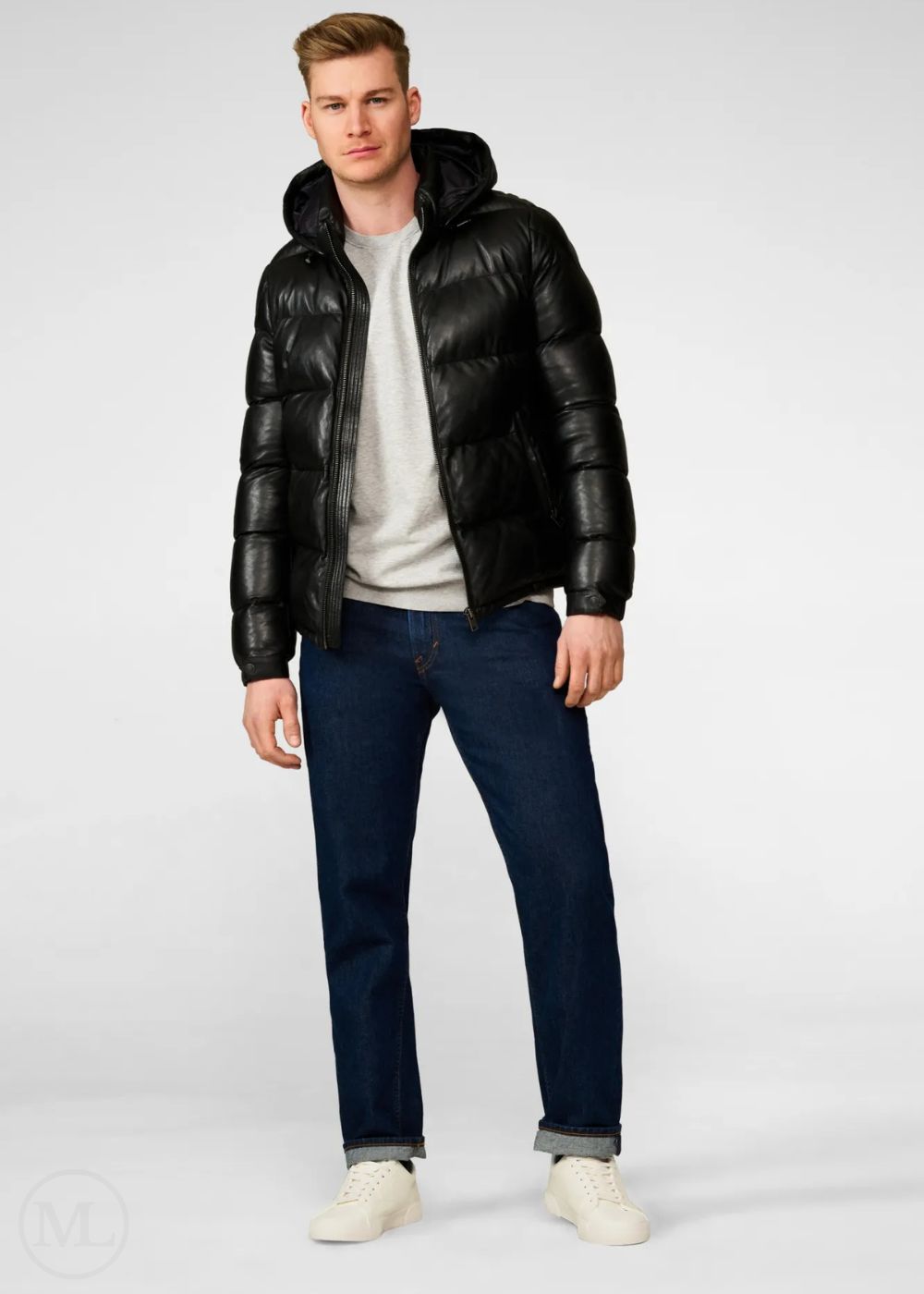 Man wearing a black leather puffer jacket, white shirt, dark jeans, and white sneakers on a white background