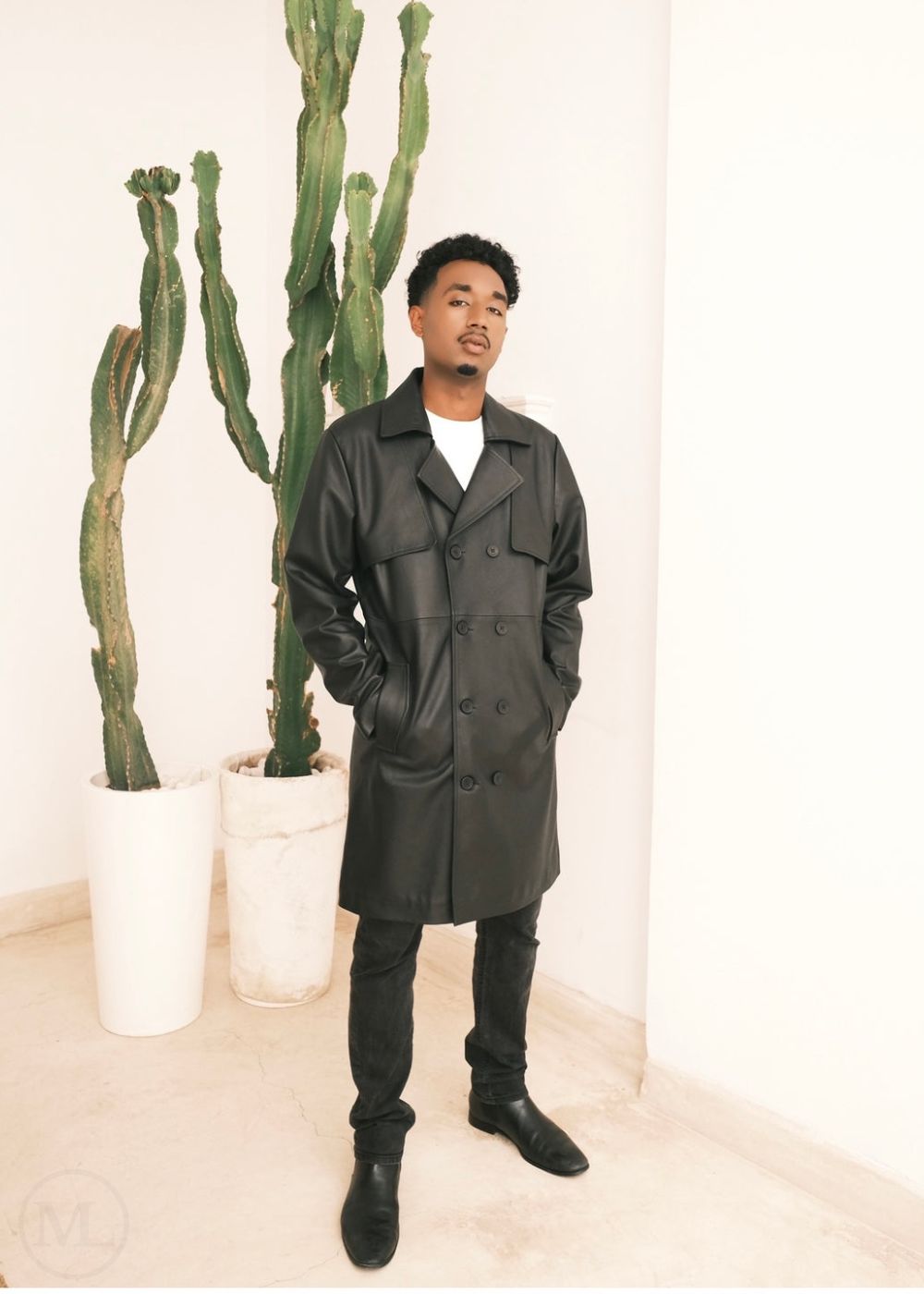 Man wearing a black leather trench coat standing in front of cacti on a light background