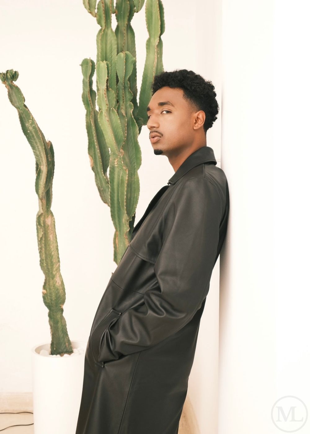 Man in a black leather trench coat standing next to a cactus plant indoors