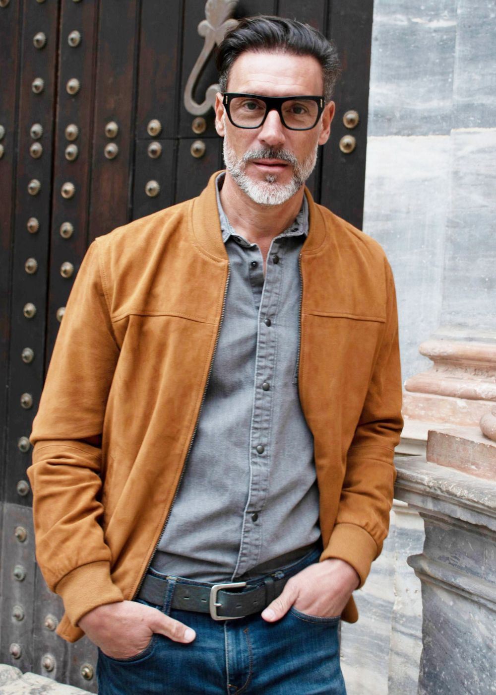 Man wearing a cognac brown suede jacket and glasses standing in front of a textured wall.