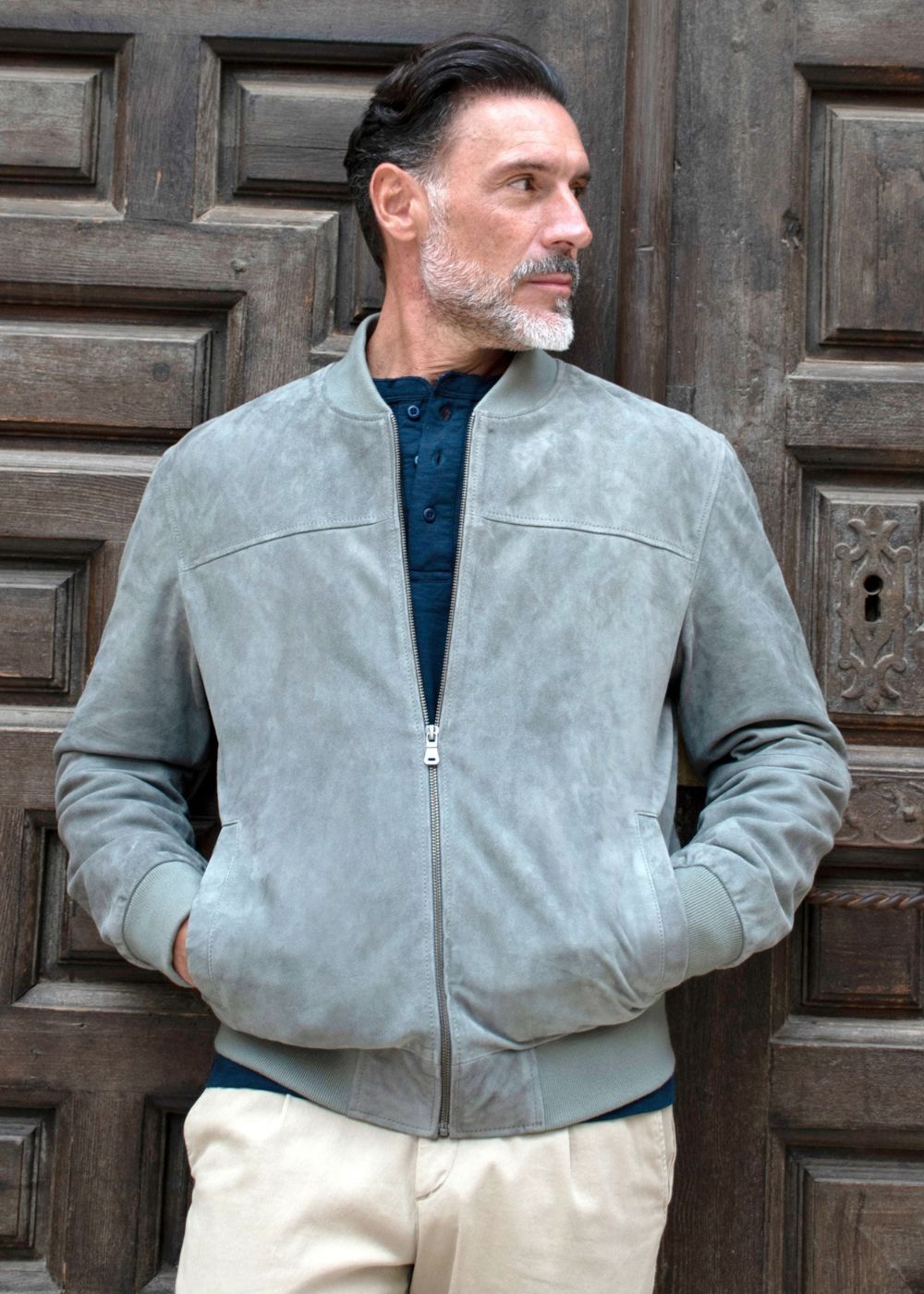 Homme portant une veste en daim gris clair debout contre une porte en bois.