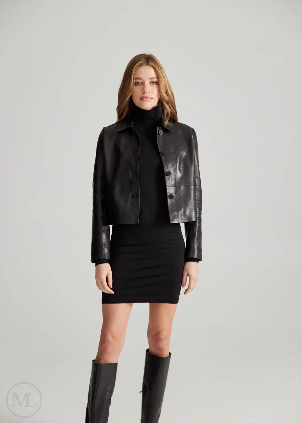 Femme portant une veste courte en cuir noir, un col roulé noir et une jupe noire avec des bottes hauteur genou sur fond gris.