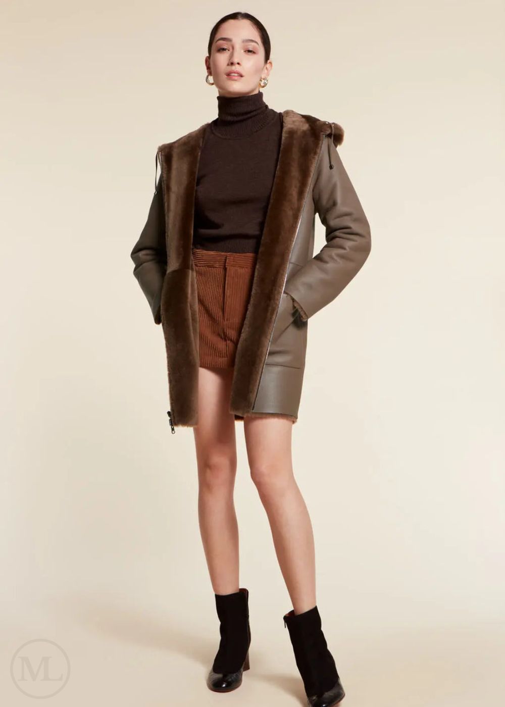 Woman wearing a brown shearling wool coat, turtleneck, shorts, and boots on a beige background
