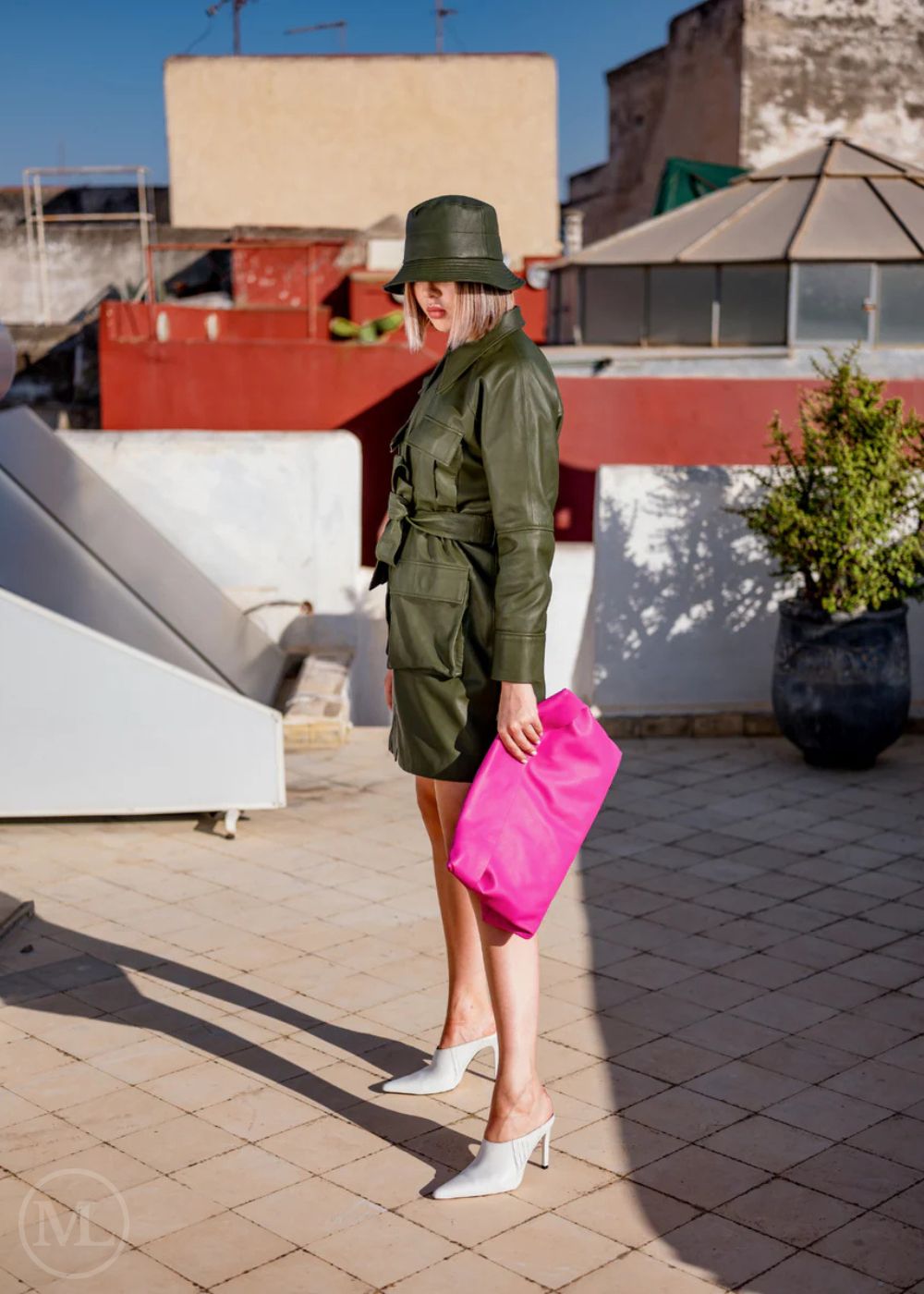 Woman wearing a Olive Green military-style Leather jacket and hat holding a pink clutch on a rooftop.