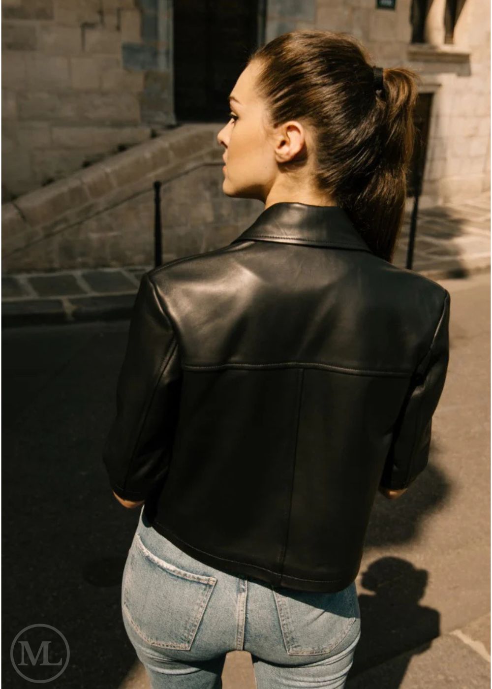 Women wearing a cropped black leather jacket and blue jeans standing on a stone pathway. back view of jacket