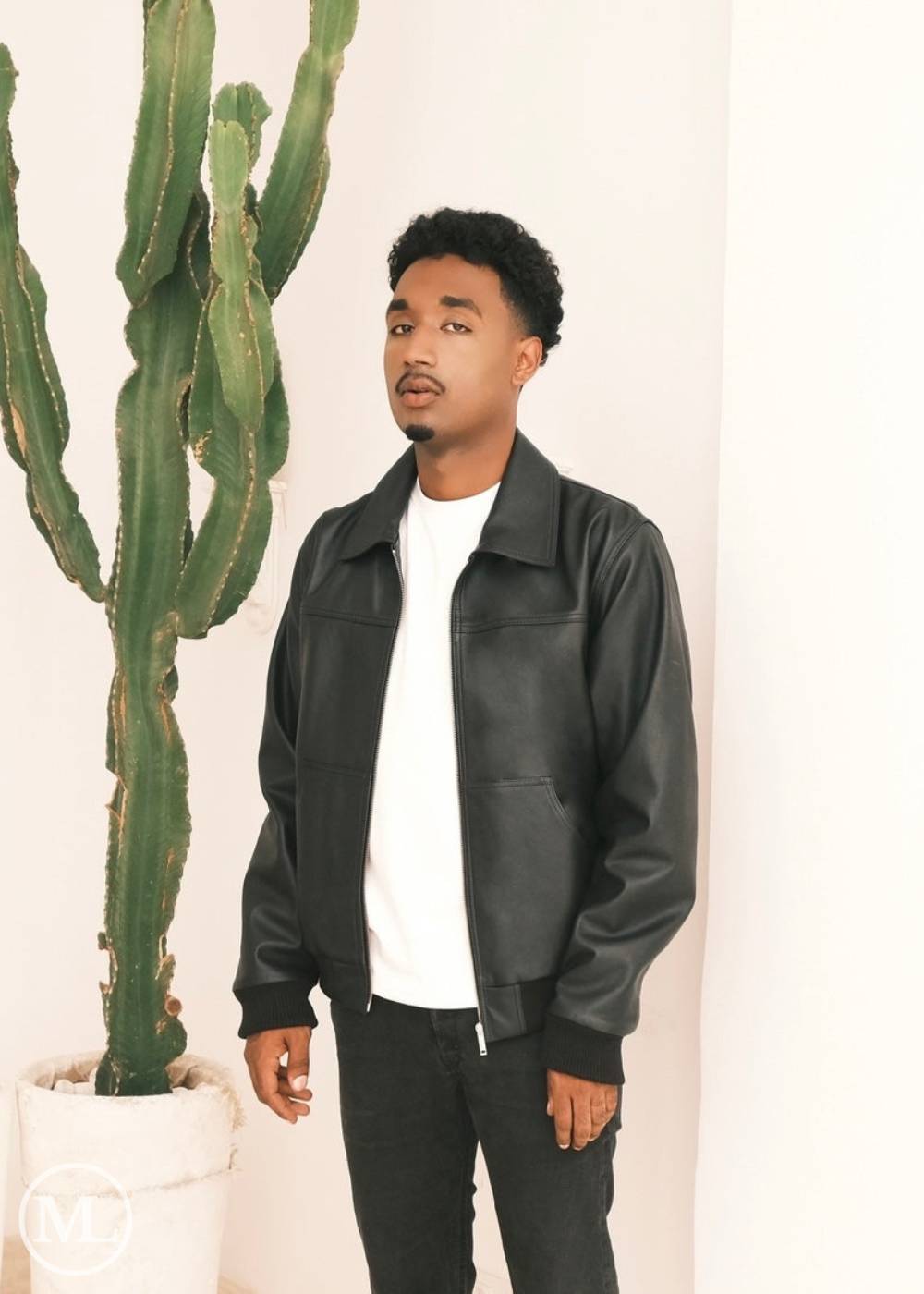 Man wearing a black leather bomber jacket with shirt collar standing next to a cactus plant indoors.
