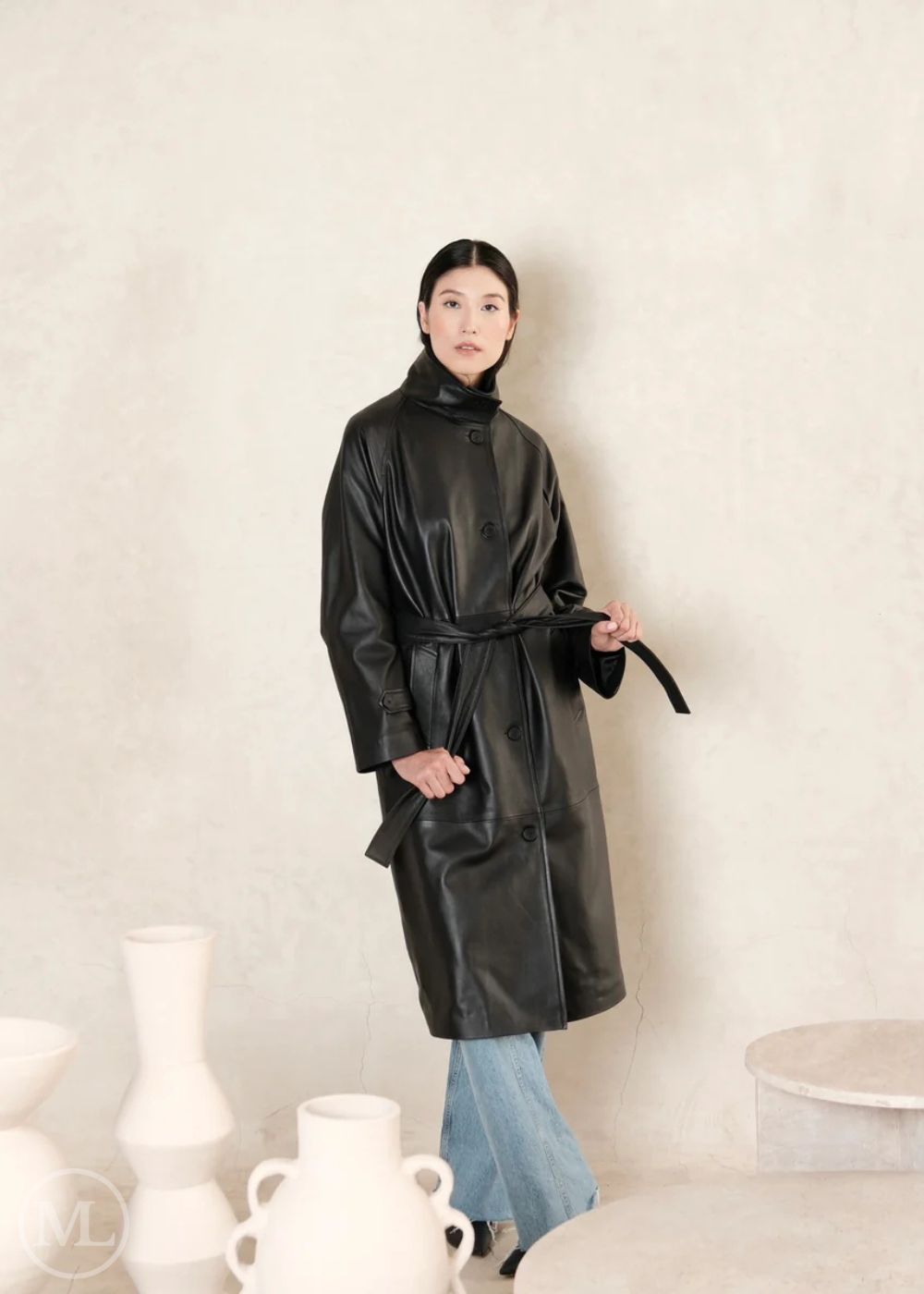women wearing a black leather trench coat holding a black belt against a beige wall