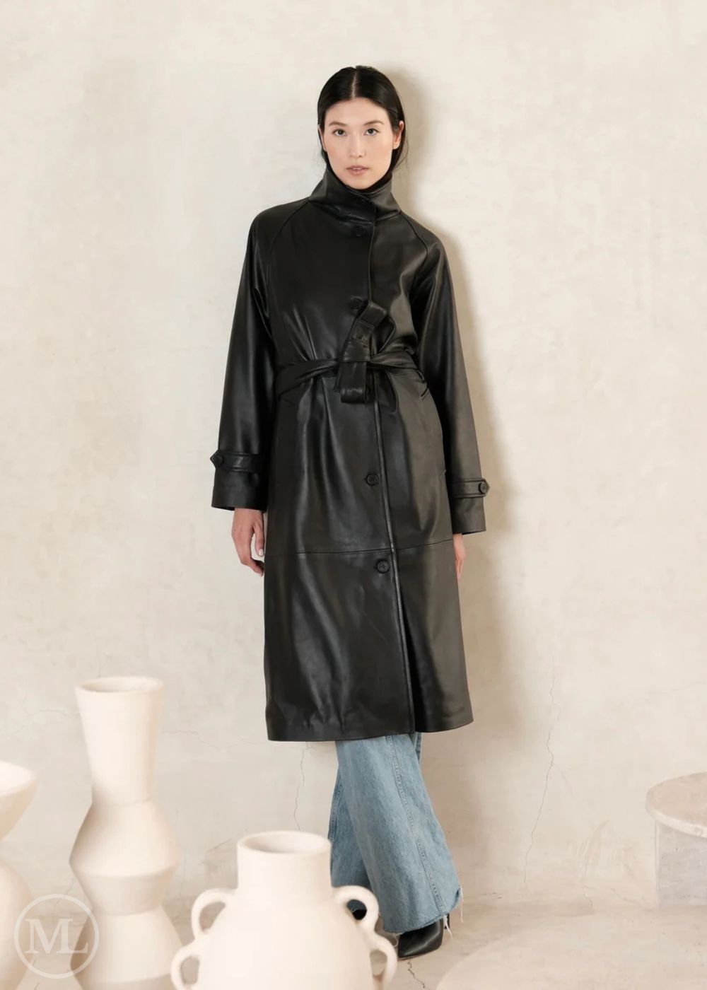 Woman wearing a black leather trench coat standing against a light background with decorative vases.