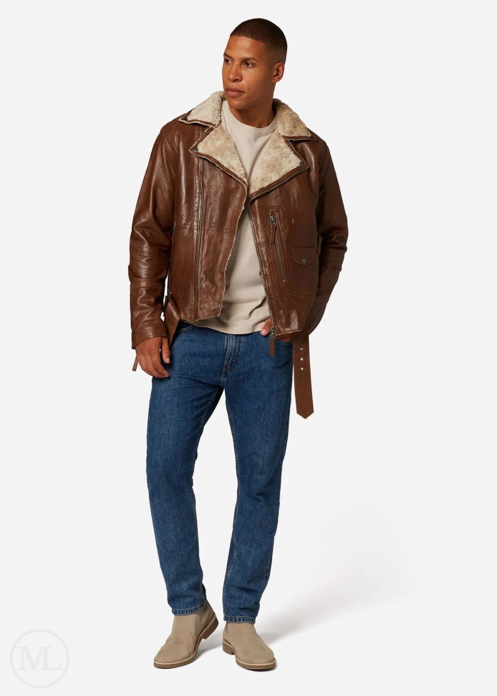 Man wearing a cognac brown leather jacket with shearling collar, white shirt, blue jeans, and beige shoes on a white background.