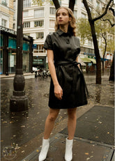 Woman in a black leather dress standing on a city street with buildings and trees in the background.