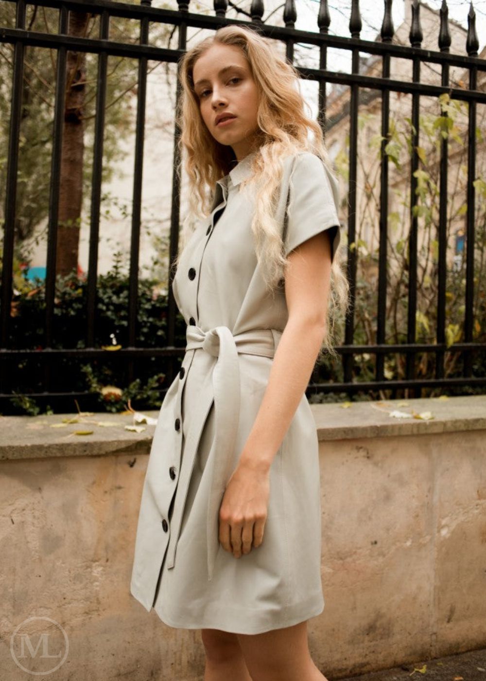 Femme vêtue d'une robe en cuir d'agneau beige pierre debout devant une clôture en métal noir.