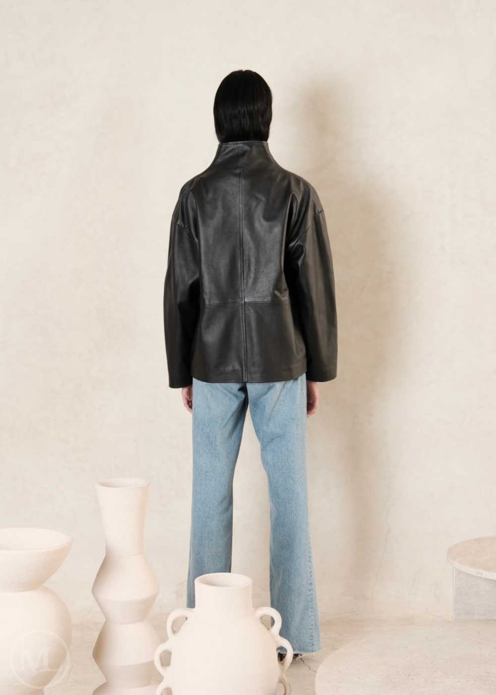 Women wearing a black leather coat and blue jeans standing in front of white vases.