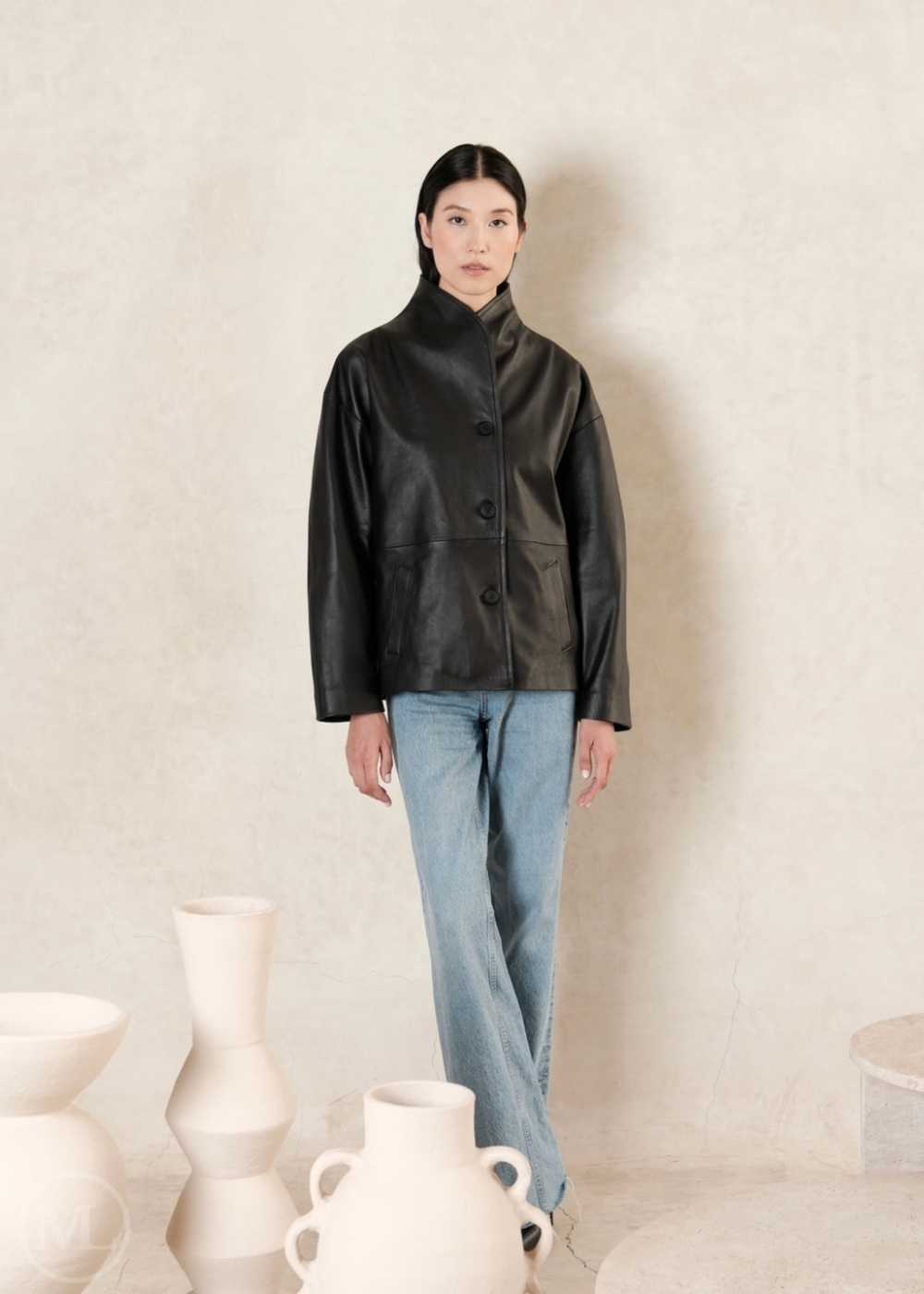 Women wearing a black leather coat style jacket and blue jeans standing against a plain background with white vases.