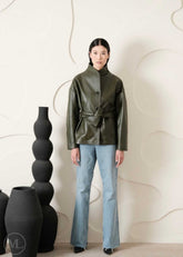 Women wearing a green leather coat style jacket and blue jeans standing against a white wall with decorative vases.