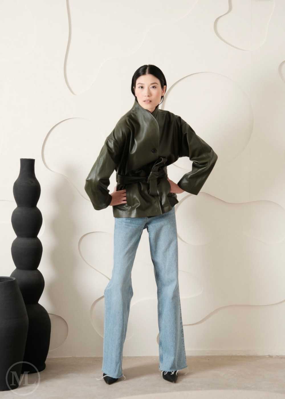 women wearing a green leather coat style jacket and light blue jeans standing against a white wall with decorative elements and commercial posing.