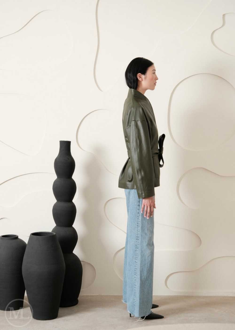 Women wearing a leather coat standing sideways, next to a series of black vases against a light wall.