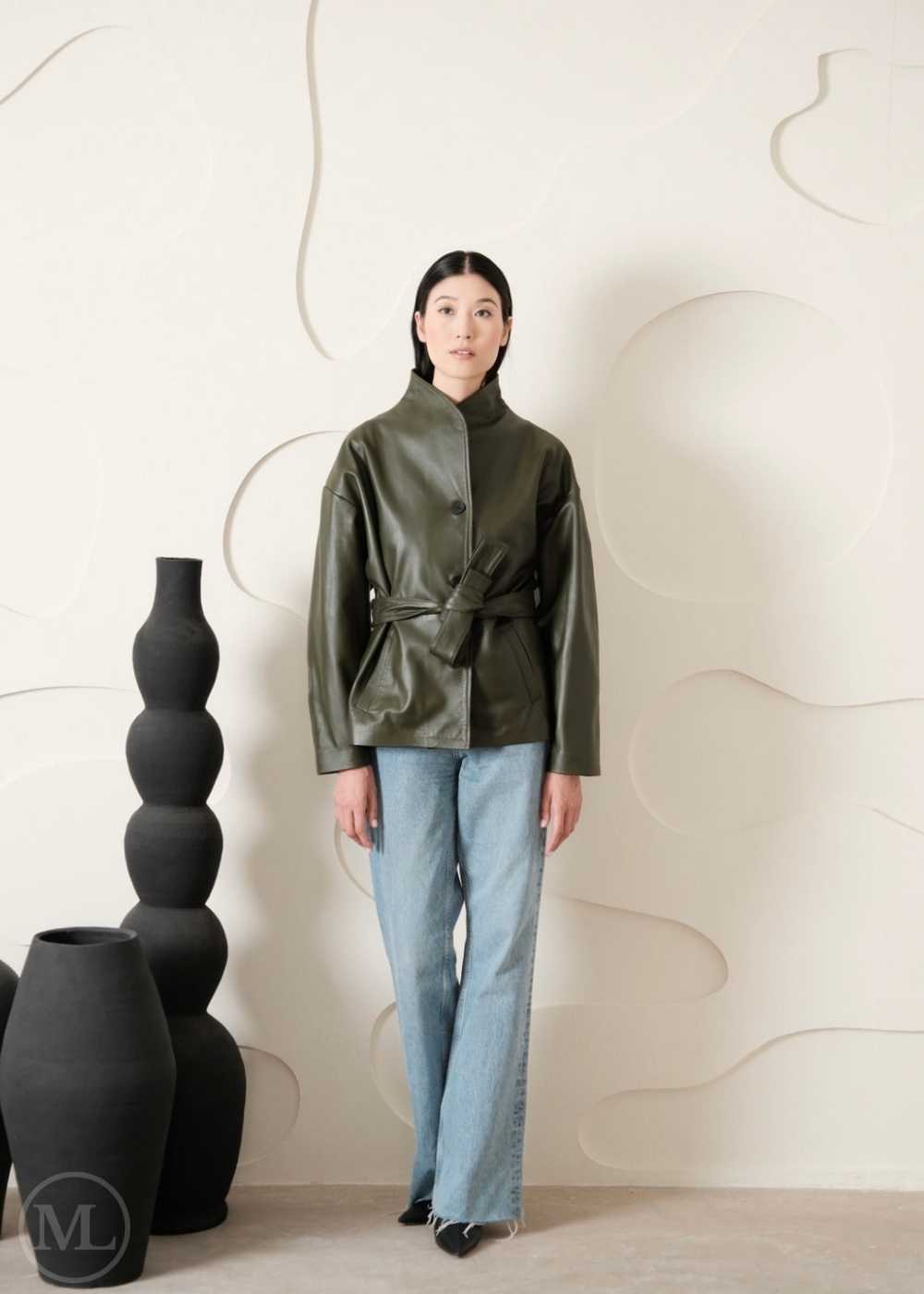 Women wearing a green leather coat style jacket and light blue jeans standing against a neutral background with decorative vases.