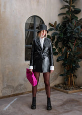 Women wearing a black leather blazer coat outfit with a hat and pink bag indoors.