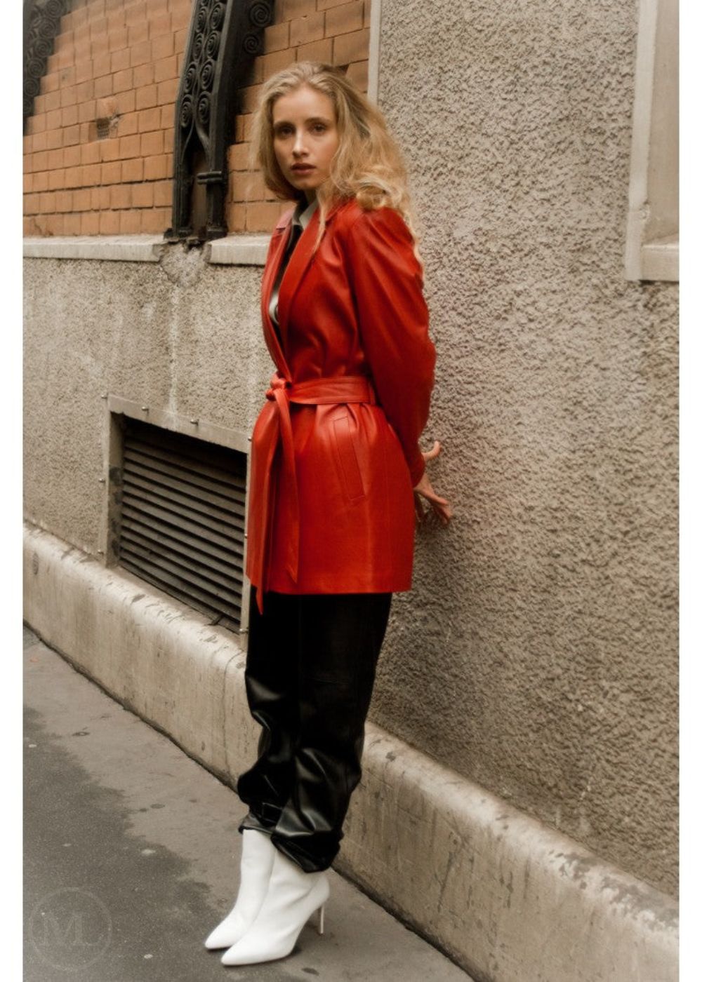 Woman in a red leather blazer coat and black pants standing against a textured wall.