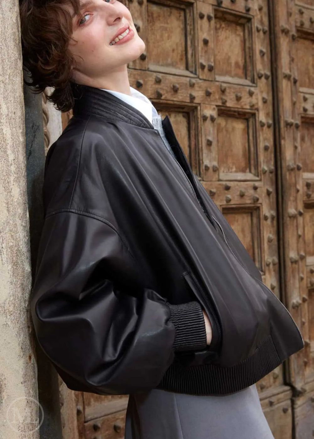 Woman wearing a black leather bomber jacket standing against a wooden door.